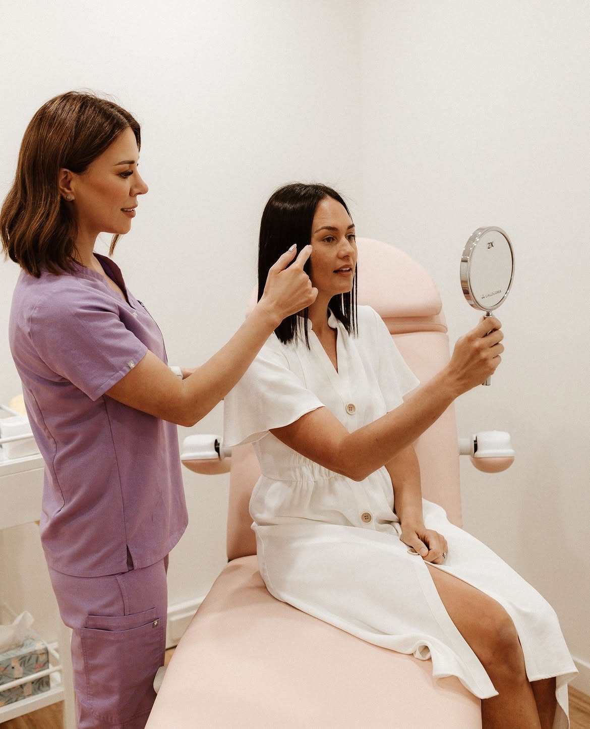 Nurse assessing a client's face while she holds a mirror in a treatment room